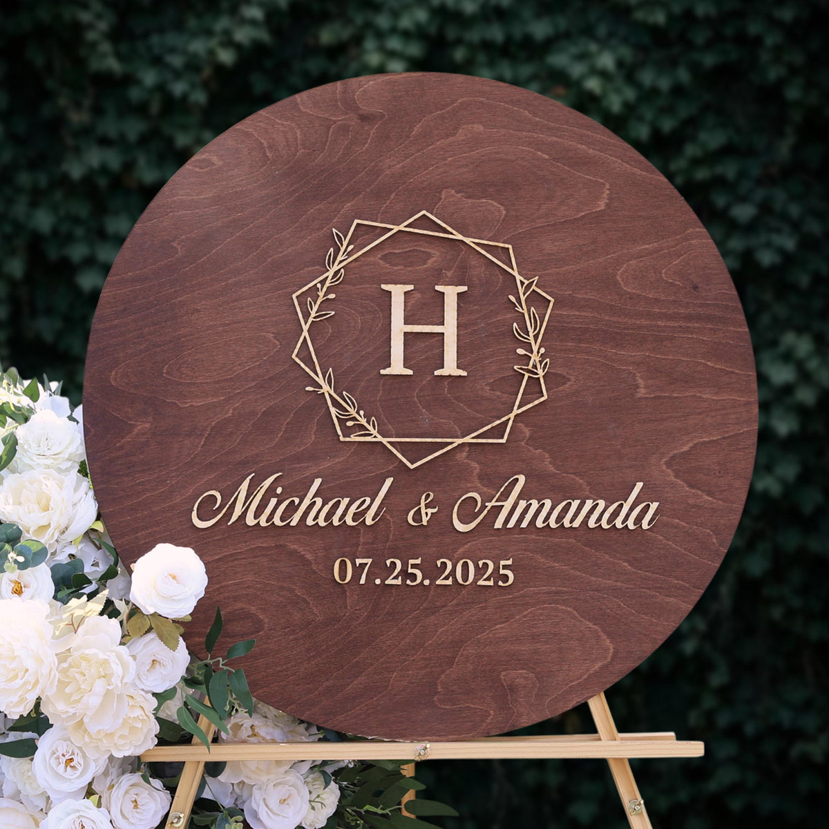 A wooden sign with the initials "H" and the names "Michael & Amanda" written on it, along with the date "07.25.2025". The sign is placed next to a bouquet of white roses.