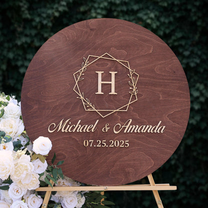 A wooden sign with the initials "H" and the names "Michael & Amanda" written on it, along with the date "07.25.2025". The sign is placed next to a bouquet of white roses.