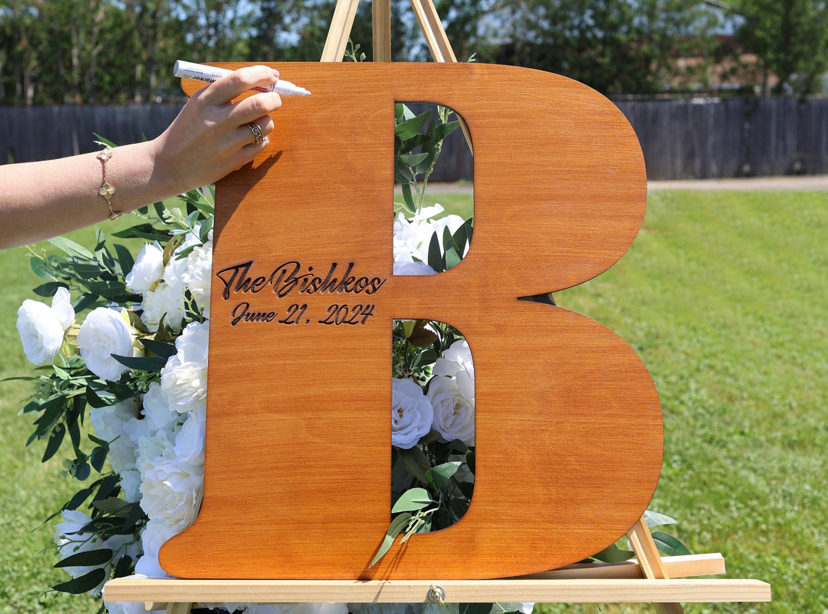 A person's hand is writing on a wooden letter B, which is surrounded by a bouquet of white flowers.