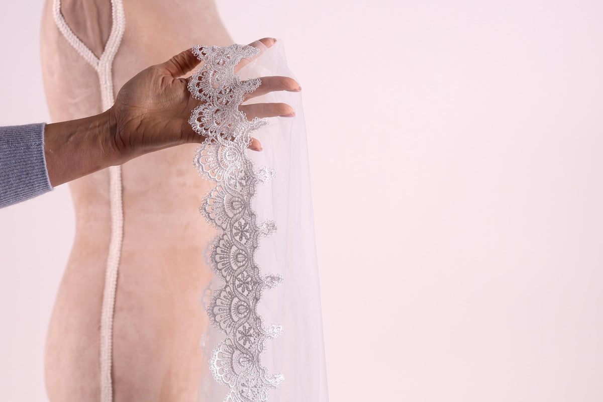 A hand holding a delicate white lace trim on a fabric, with a blurred background.