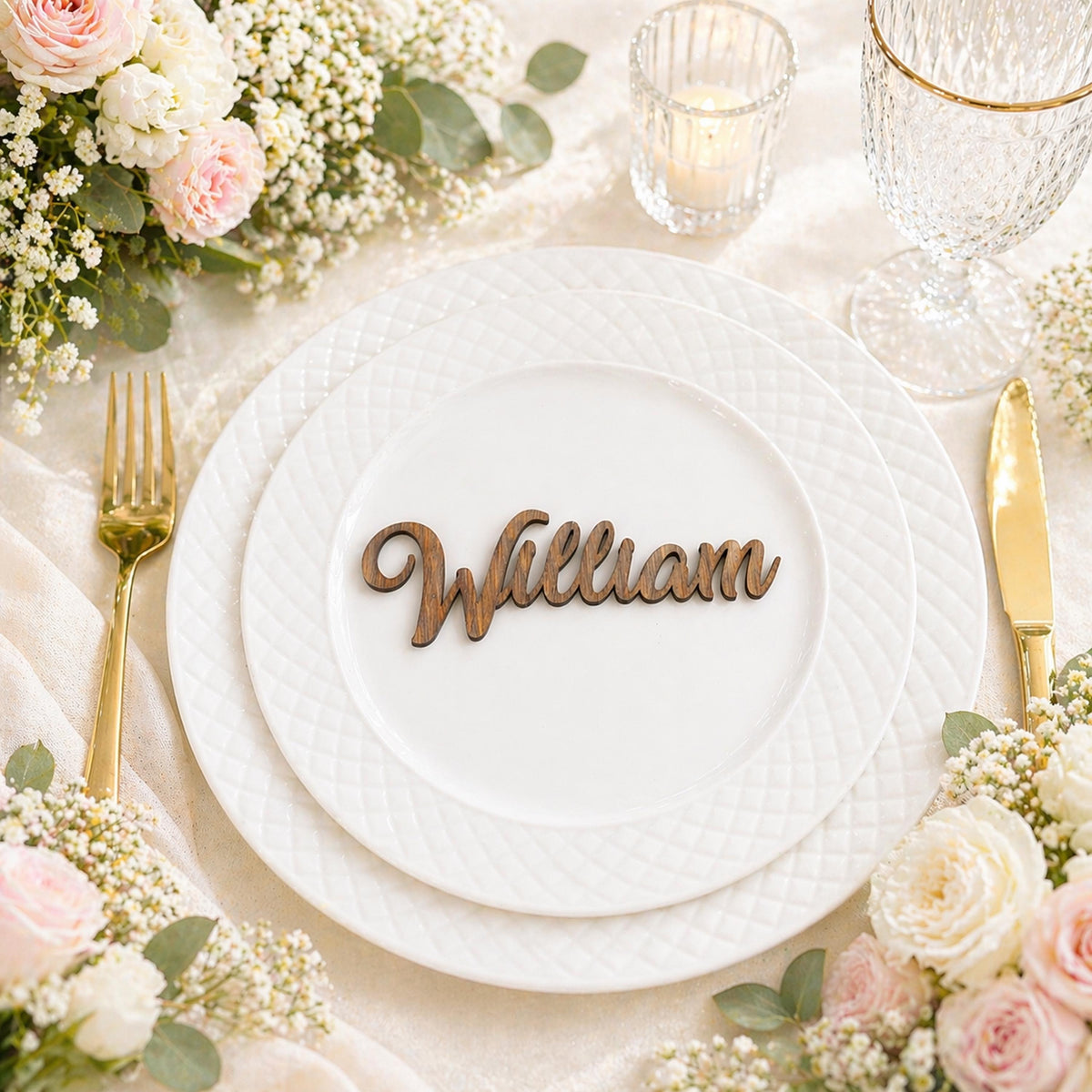 A table setting with a white plate, goldware, and a wooden nameplate with the name "William" on it. The table is decorated with white flowers and greenery, creating a formal and elegant atmosphere.
