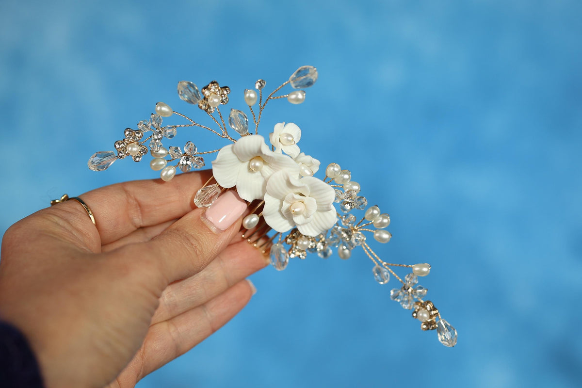 A hand holding a decorative hair accessory with white flowers and clear gemstones against a blue background.