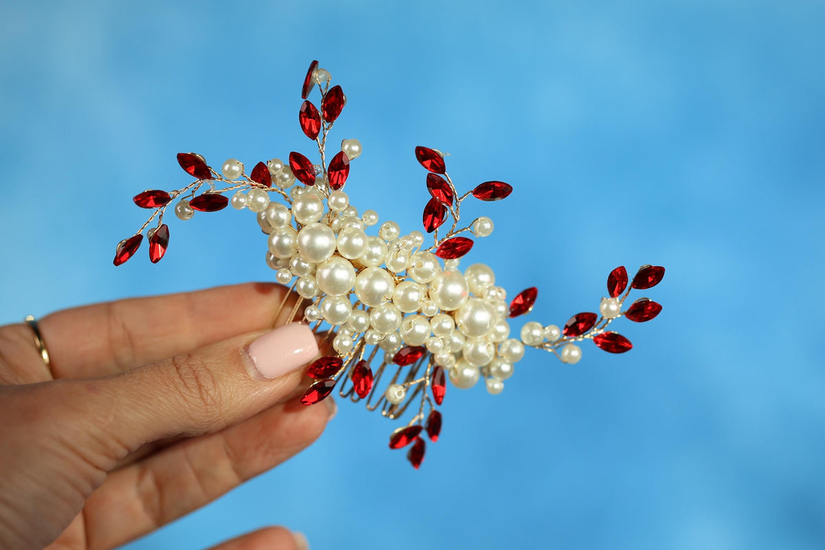 A hand holding a decorative hair accessory with red and white elements, including pearls and gemstones, against a blue background.