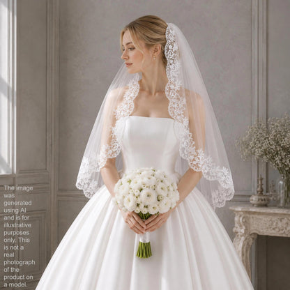 A woman in a white wedding dress holding a bouquet of white flowers stands in a room with a window and a table with flowers.