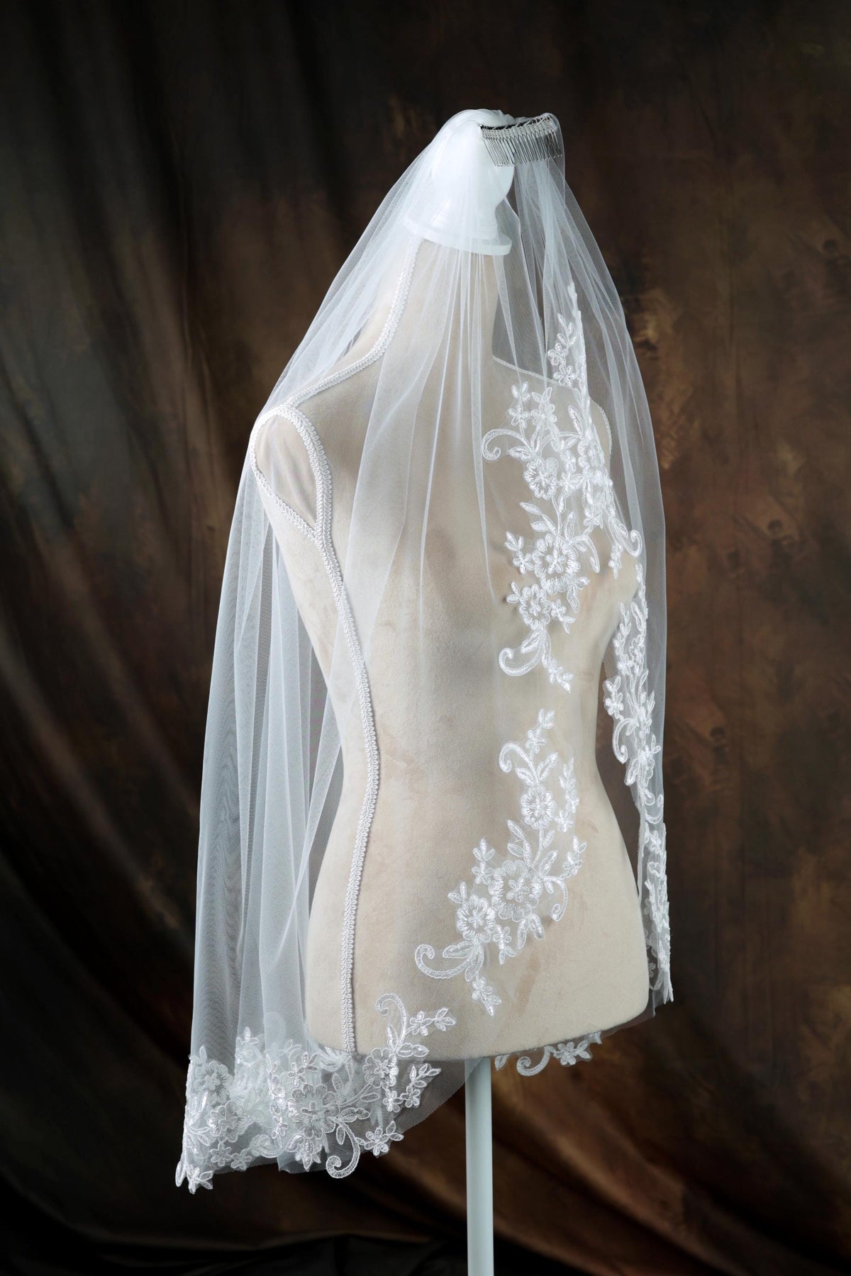 A white veil with a floral pattern is displayed on a mannequin against a dark background.