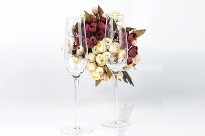 Two wine glasses with personalized messages stand in front of a bouquet of red and white roses.