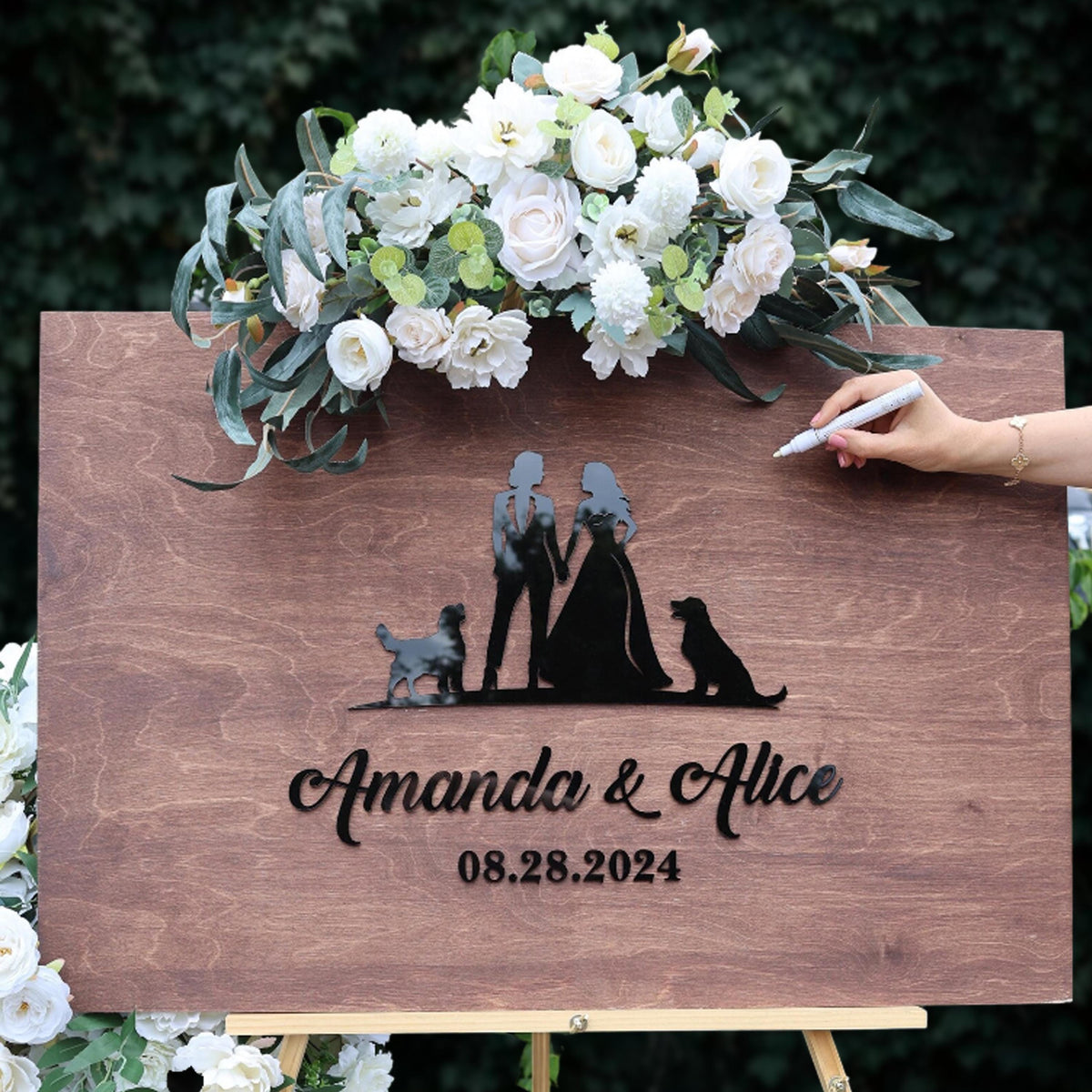 a wooden sign with a floral arrangement and a person's silhouette of a couple, along with the names 'Amanda & Alice' and the date '08.28.2024'.