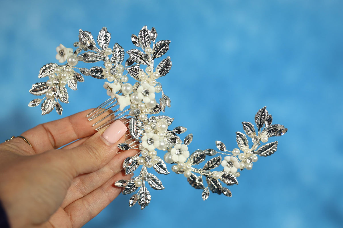 A hand holding a decorative hair accessory with white flowers and leaves against a blue background.