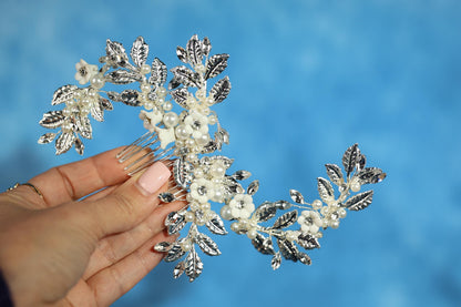 A hand holding a decorative hair accessory with white flowers and leaves against a blue background.