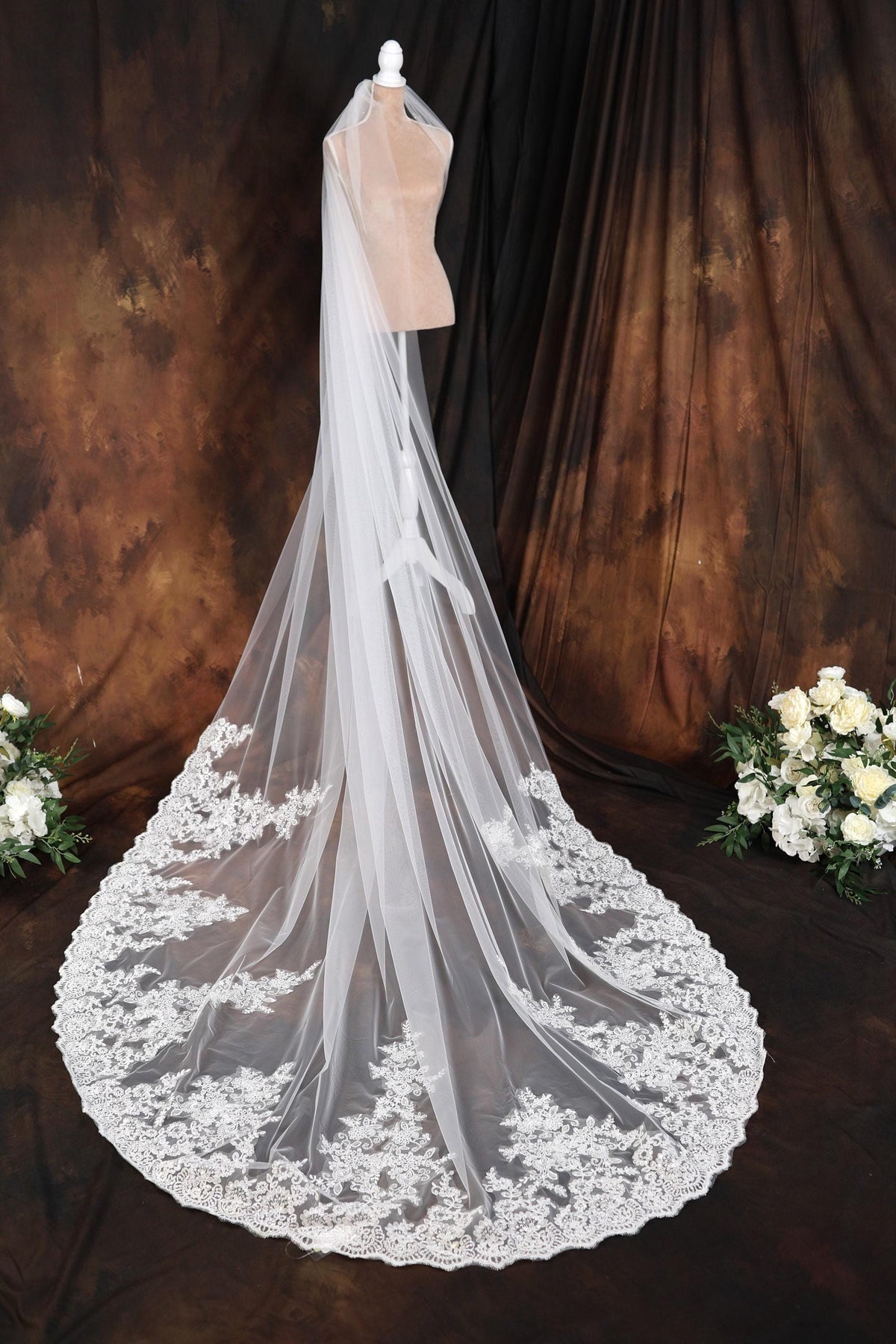 A white wedding veil with a long train and lace trim, displayed on a mannequin head against a brown background with white flowers.