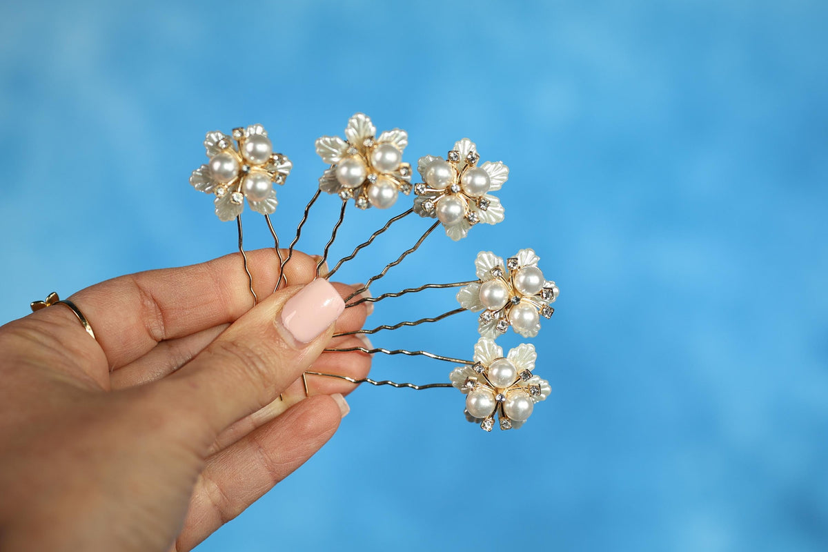 A hand holding a group of six silver hair clips with white flowers attached to them against a blue background.