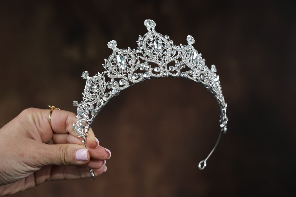 A hand holding a silver tiara with intricate designs and sparkling gemstones against a dark background.