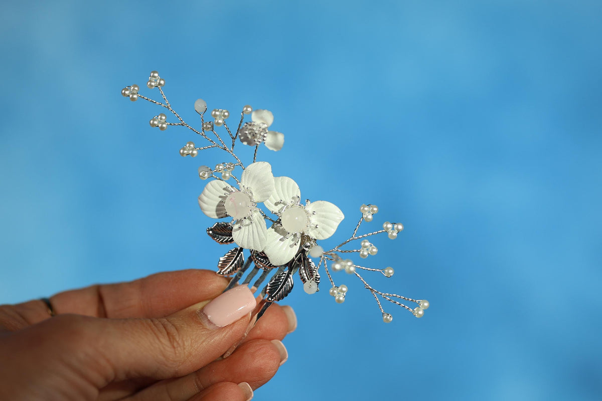 A hand holding a decorative hair clip with white flowers and silver beads against a blue background.