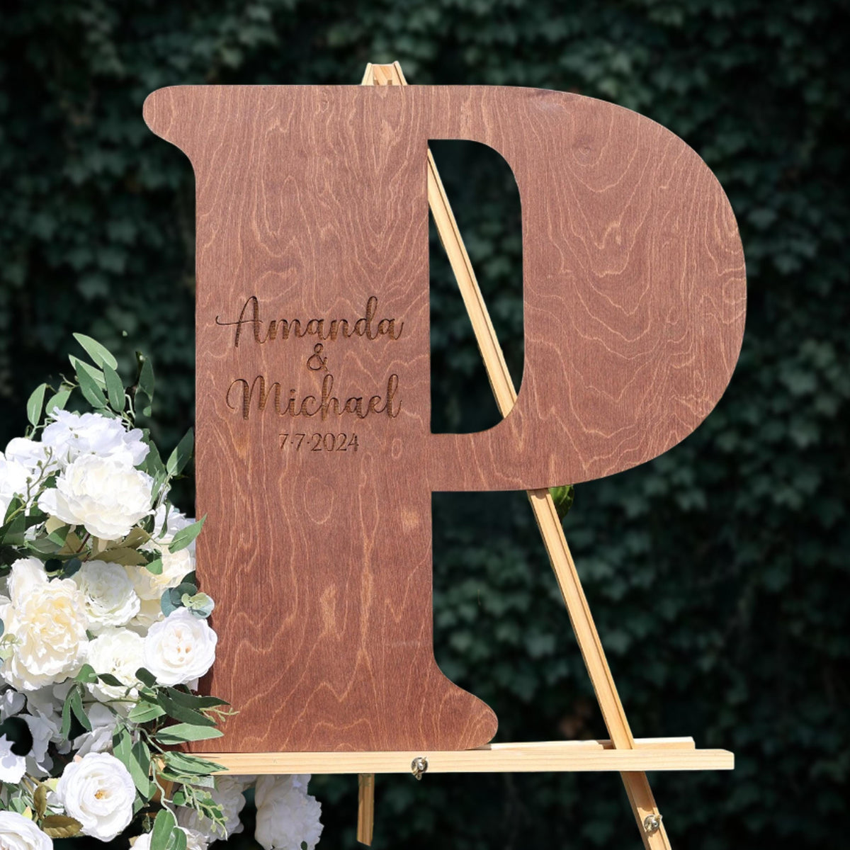 A wooden letter 'P' with the names 'Amanda & Michael' engraved on it, standing on a wooden easel with white flowers.
