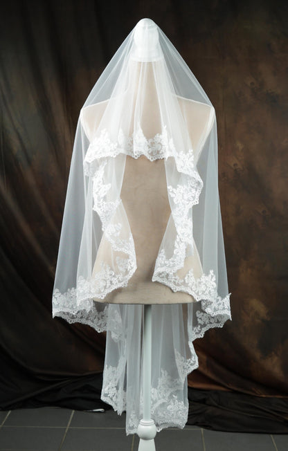 A white wedding veil with intricate lace detailing is displayed on a mannequin against a dark background.