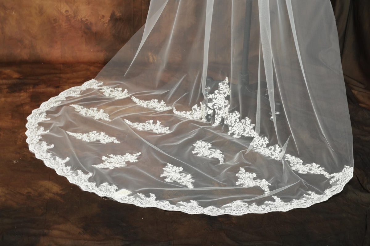 A close-up view of a delicate white lace veil with intricate floral embroidery, displayed on a brown background.