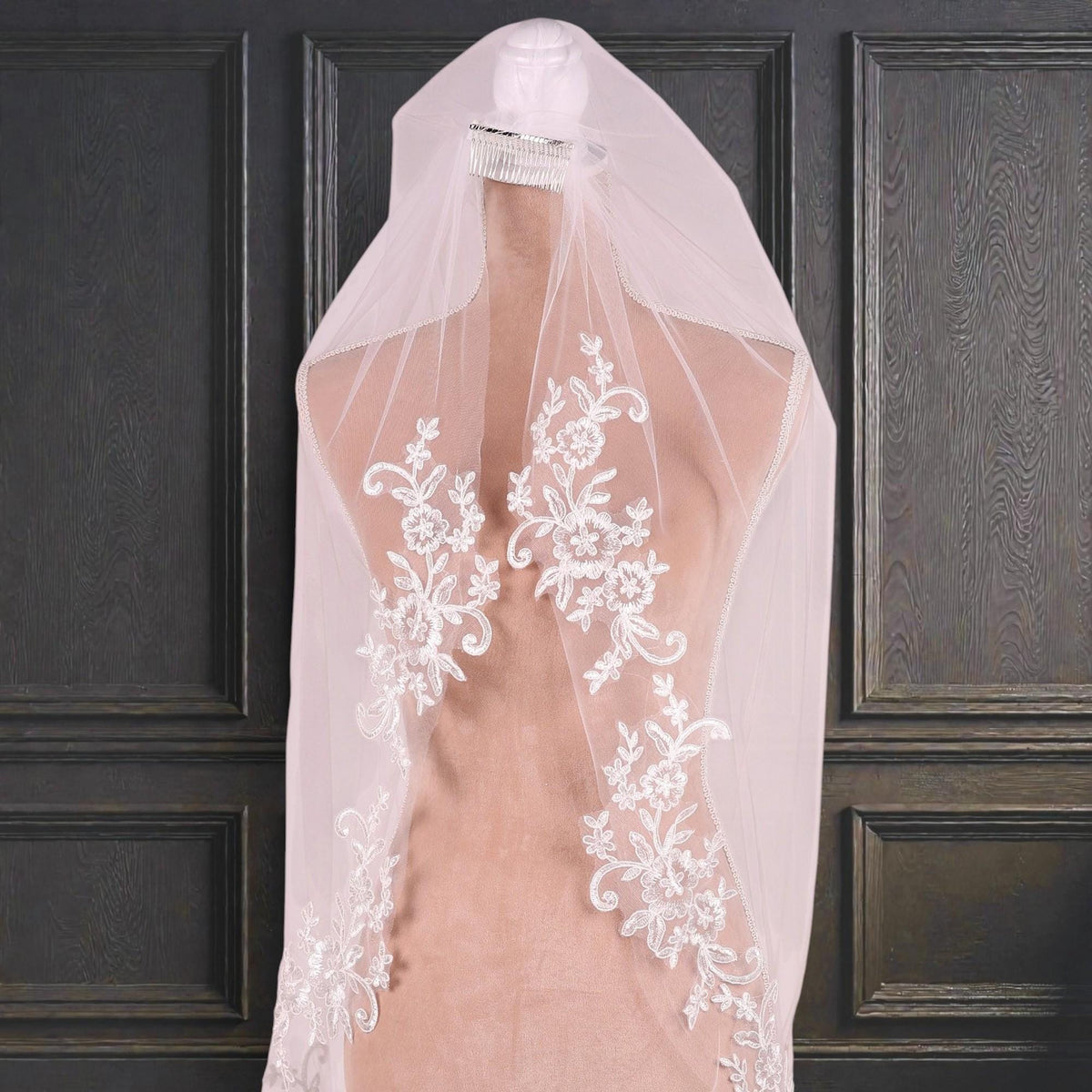 A white wedding veil with intricate floral embroidery is displayed on a mannequin against a dark wooden panel background.