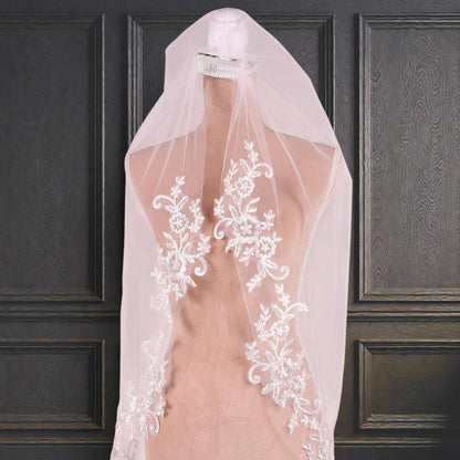 A white wedding veil with intricate floral embroidery is displayed on a mannequin against a dark wooden panel background.