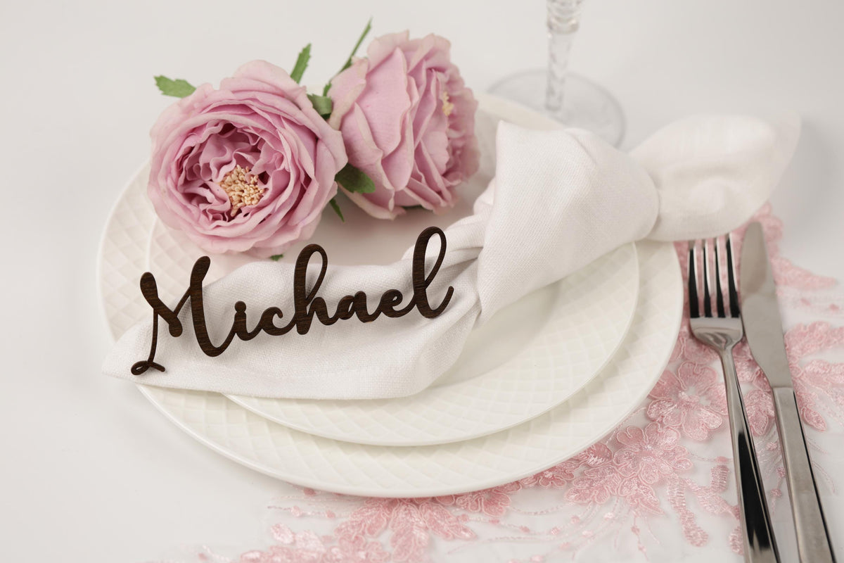 A table setting with a white plate, a napkin with the name "Michael" written on it, and two pink roses. The table also has silverware, including a fork and knife, and a glass.