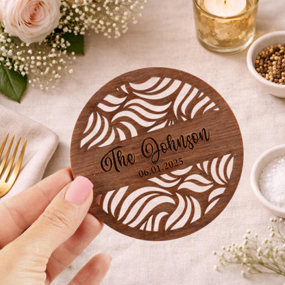 A hand holding a wooden coaster with a decorative design, surrounded by various tableware items and flowers.