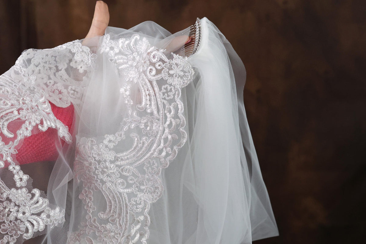 A hand holding a white veil with intricate lace and floral embroidery.