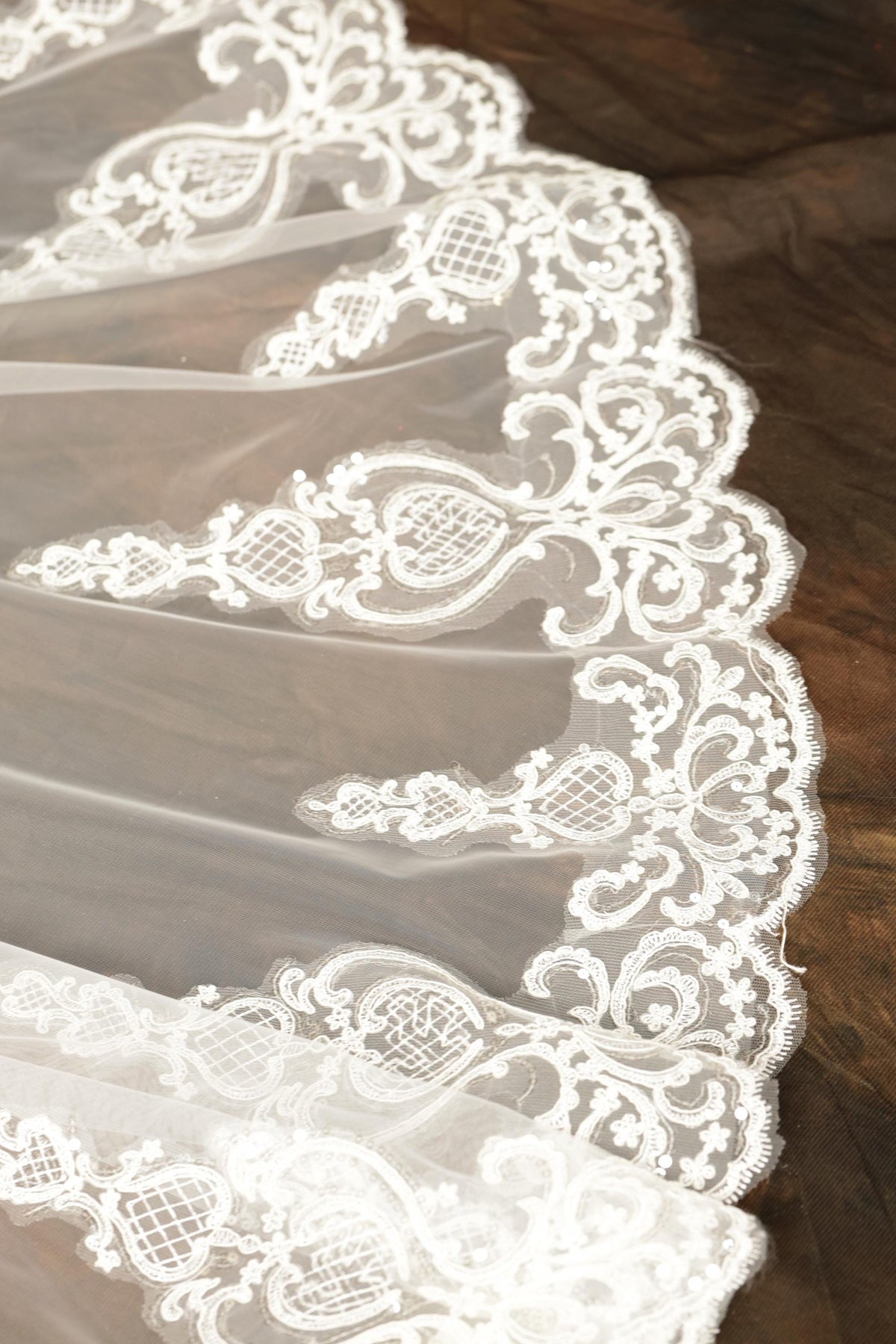 a close-up view of a delicate, white lace veil with intricate floral patterns. The veil appears to be made of sheer fabric and is displayed on a wooden surface.