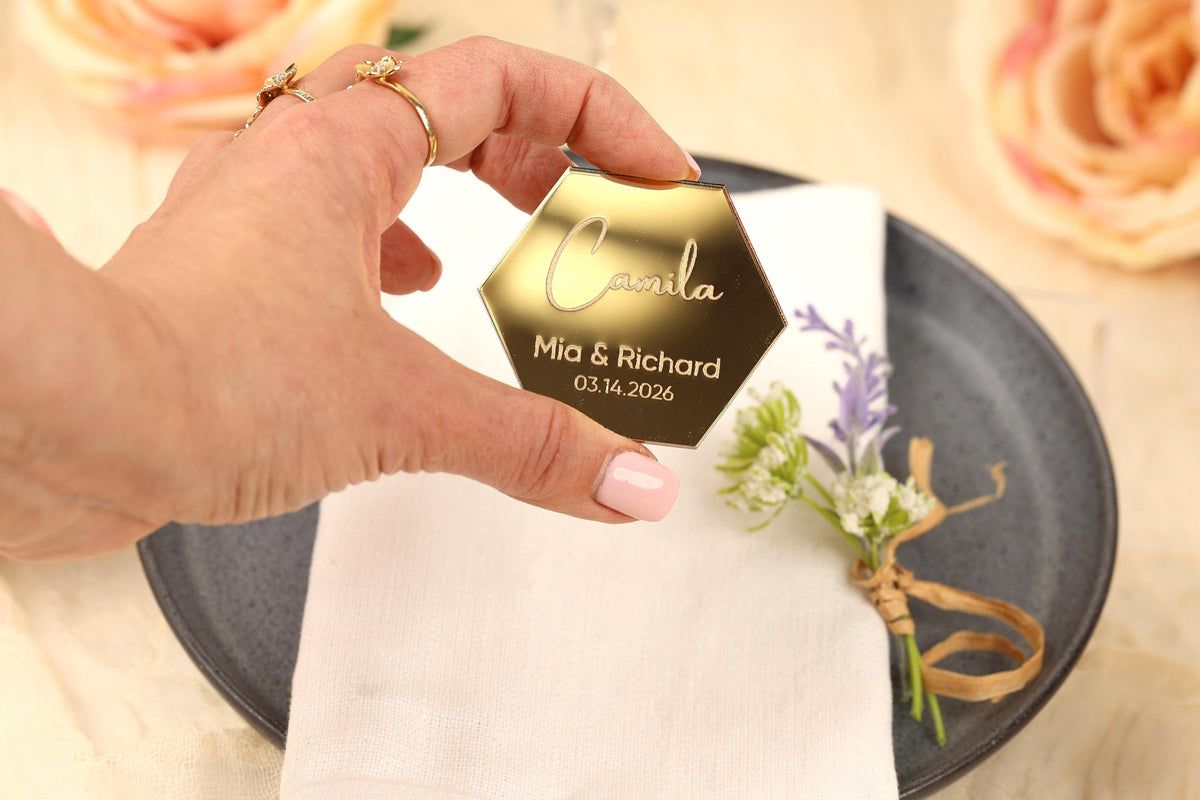 A hand holding a gold hexagonal wedding favor with the names 'Carmela' and 'Mia & Richard' engraved on it, along with the date '03.14.2026'.