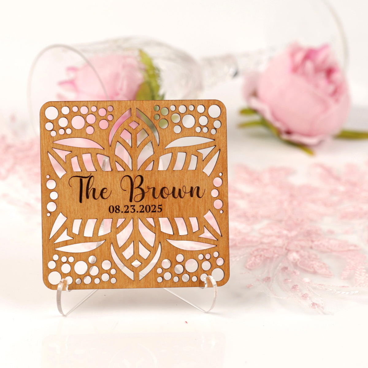 A wooden sign with a floral design and text, placed on a white surface with a pink rose in the background.