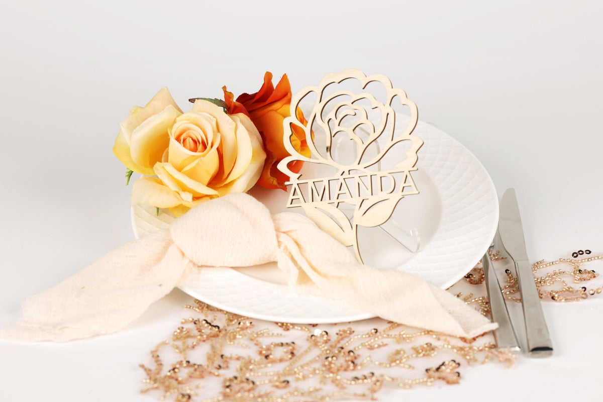 a white plate with a wooden name card, a yellow rose, and a beaded necklace, all arranged on a white surface.