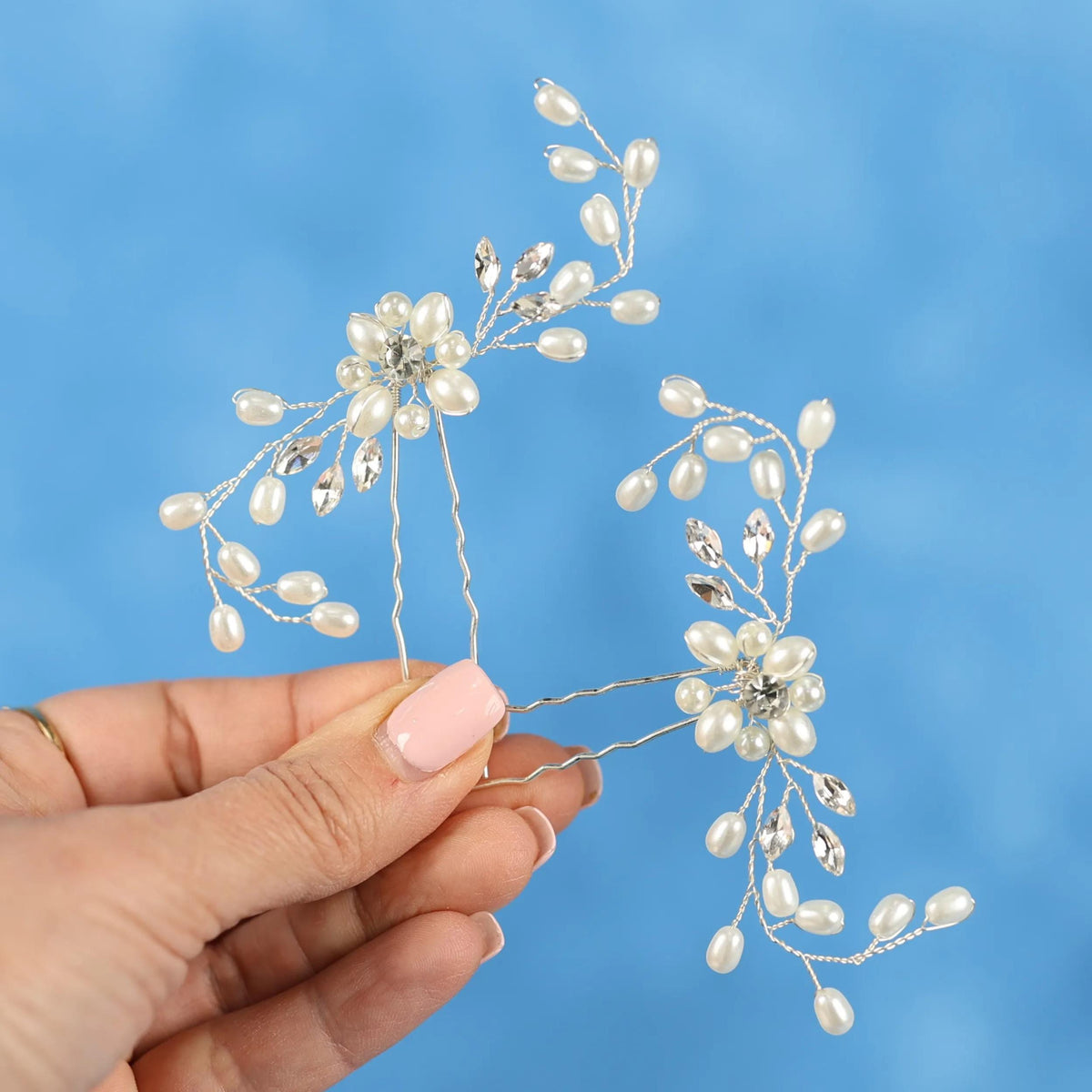 A hand holding two silver hair clips adorned with white pearls and crystals against a blue background.