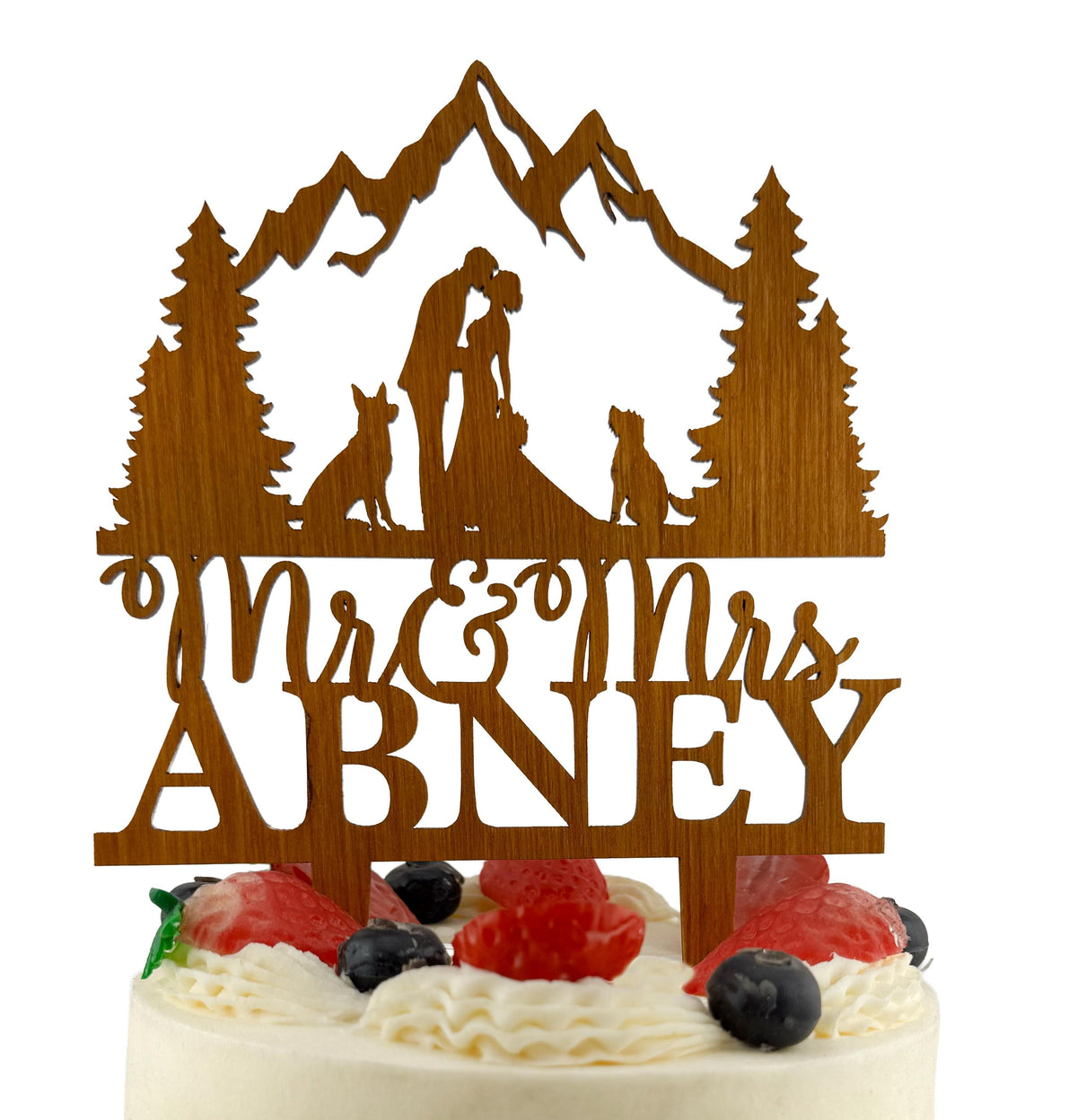 A wooden wedding cake topper with the names 'Mr & Mrs Abney' carved into it, featuring silhouettes of a couple embracing and a dog.