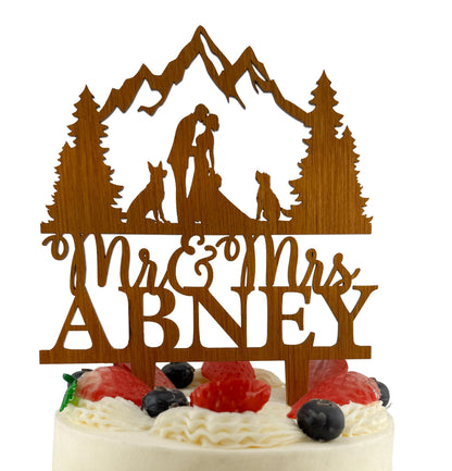 A wooden wedding cake topper with the names 'Mr & Mrs Abney' carved into it, featuring silhouettes of a couple embracing and a dog.