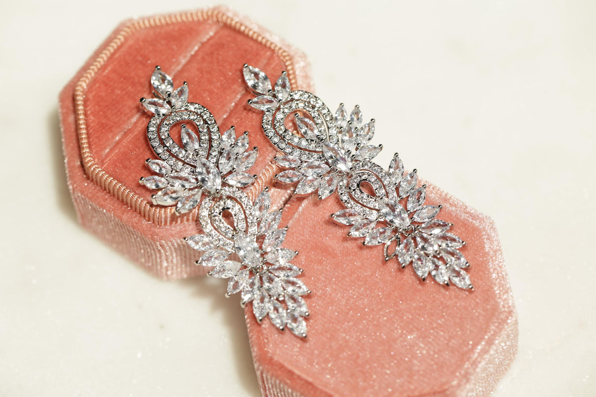 A pair of elegant diamond earrings with intricate floral designs rests on a pink velvet box.
