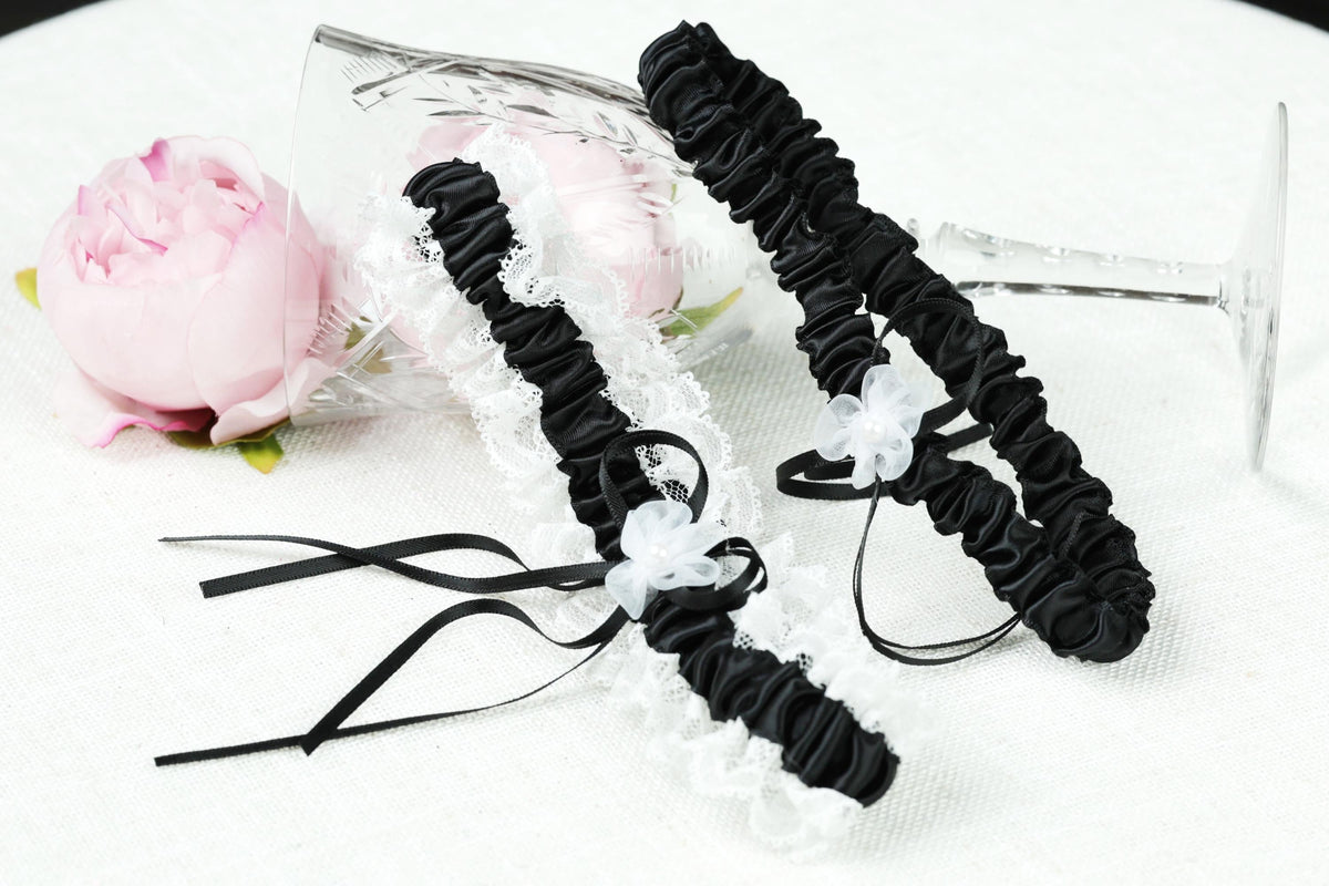 a pair of black and white lace wedding garter with black ribbons, placed on a white surface next to a pink rose.