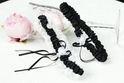a pair of black and white lace wedding garter with black ribbons, placed on a white surface next to a pink rose.