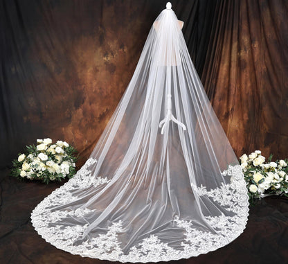 A white wedding veil with a long, flowing train and a lace border is displayed on a mannequin against a brown background.