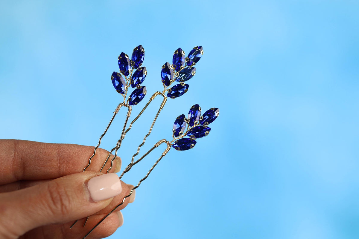 A hand holding a pair of hair clips with blue gemstone leaves against a blue background.