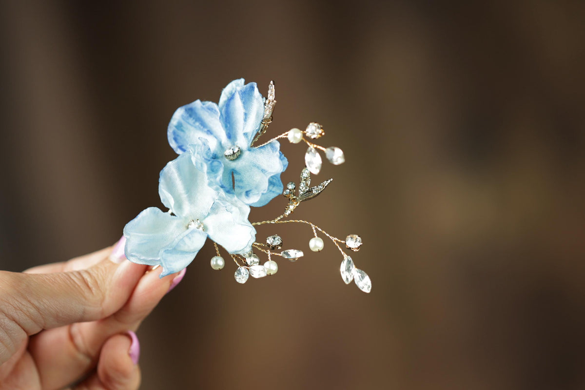 A hand holding a delicate blue flower adorned with pearls and gemstones against a blurred background.