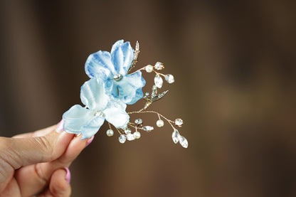 A hand holding a delicate blue flower adorned with pearls and gemstones against a blurred background.