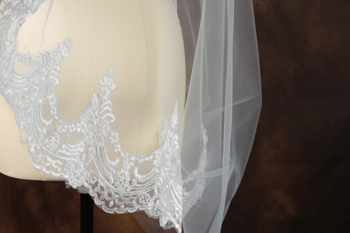 A close-up view of a delicate white lace veil with intricate embroidery, displayed on a mannequin or a similar object.