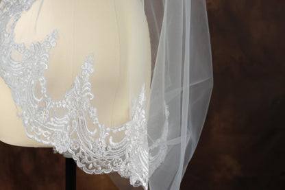 A close-up view of a delicate white lace veil with intricate embroidery, displayed on a mannequin or a similar object.