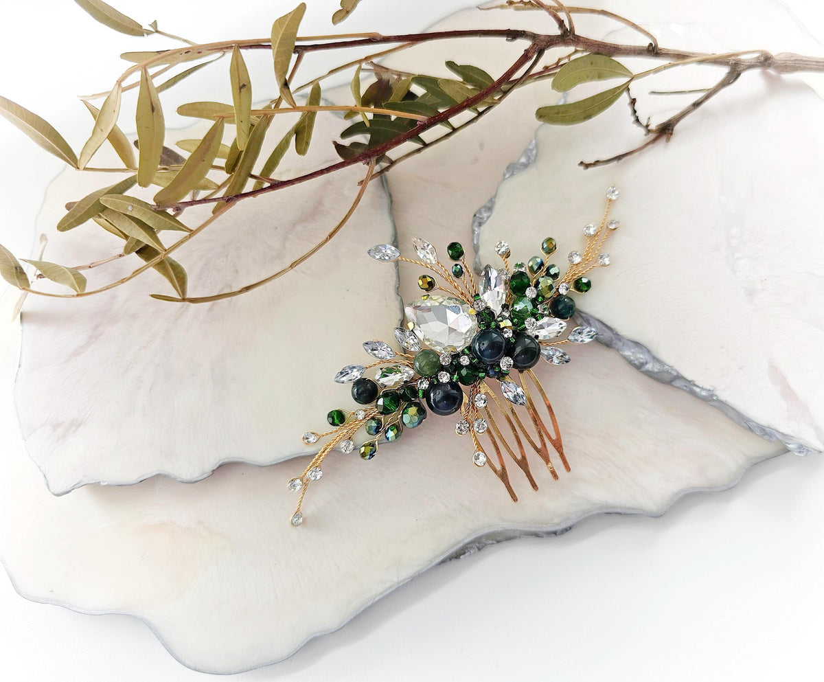 A decorative hair accessory with a gold-colored metal frame and a floral design, featuring green and white gemstones, is displayed on a white marble surface.
