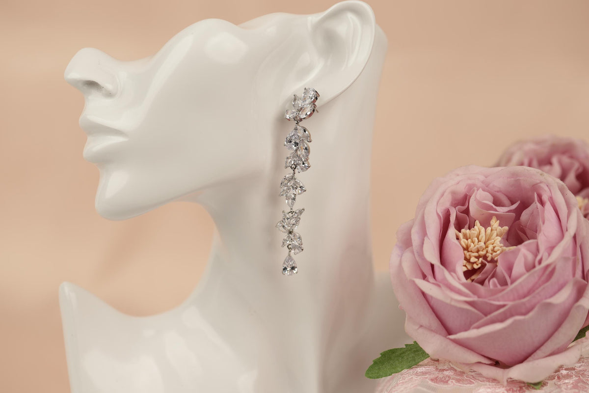 A white mannequin head wearing a pair of dangling earrings with small, clear gemstones. The earrings are placed next to a pink rose, creating a visually appealing and elegant display.
