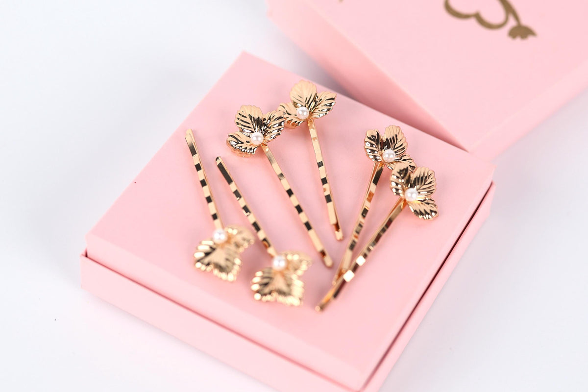 A pink box containing several gold hair clips with leaf-shaped designs.