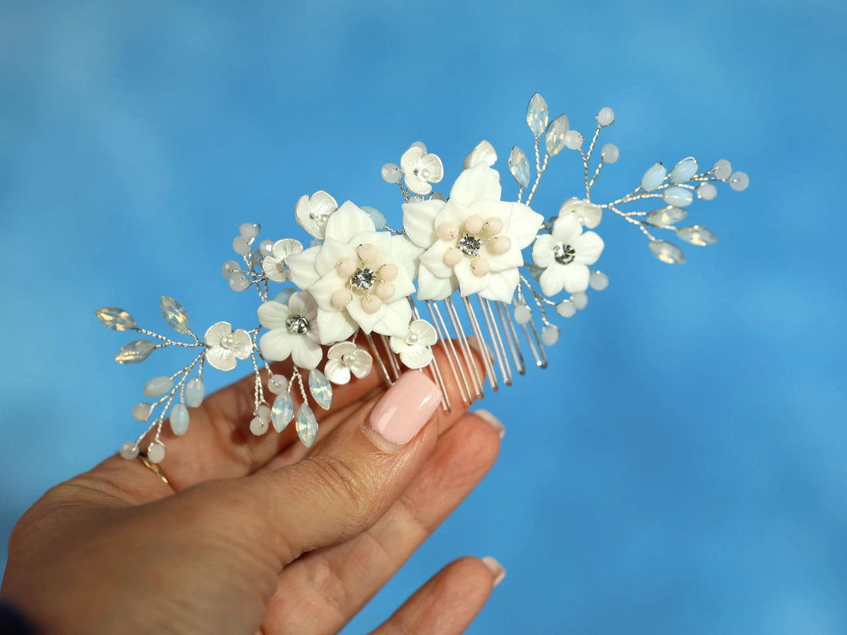 A hand holding a decorative hair accessory with white flowers and silver embellishments against a blue background.