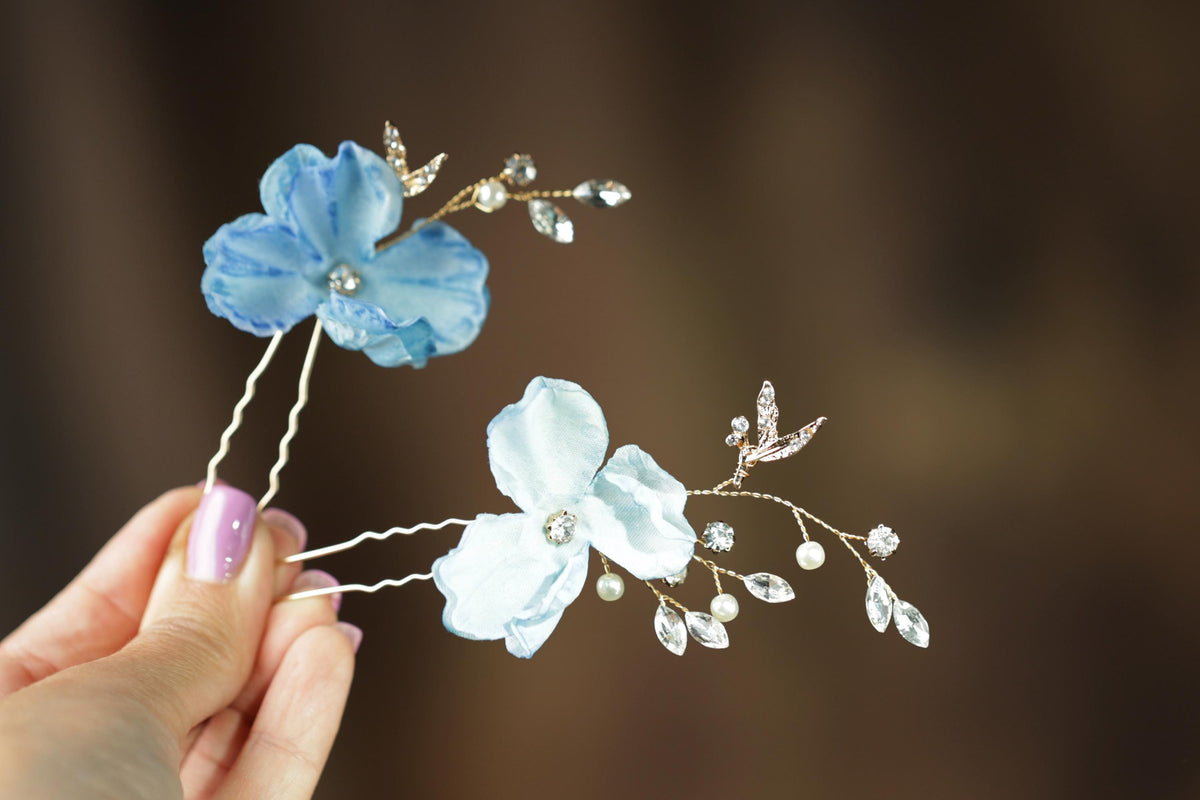 A hand holding two delicate flower-shaped hair clips with blue and white petals and sparkling rhinestones.