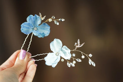 A hand holding two delicate flower-shaped hair clips with blue and white petals and sparkling rhinestones.