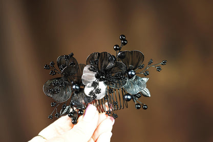 A hand holding a decorative hair accessory with black and silver metallic elements, including flowers and berries.
