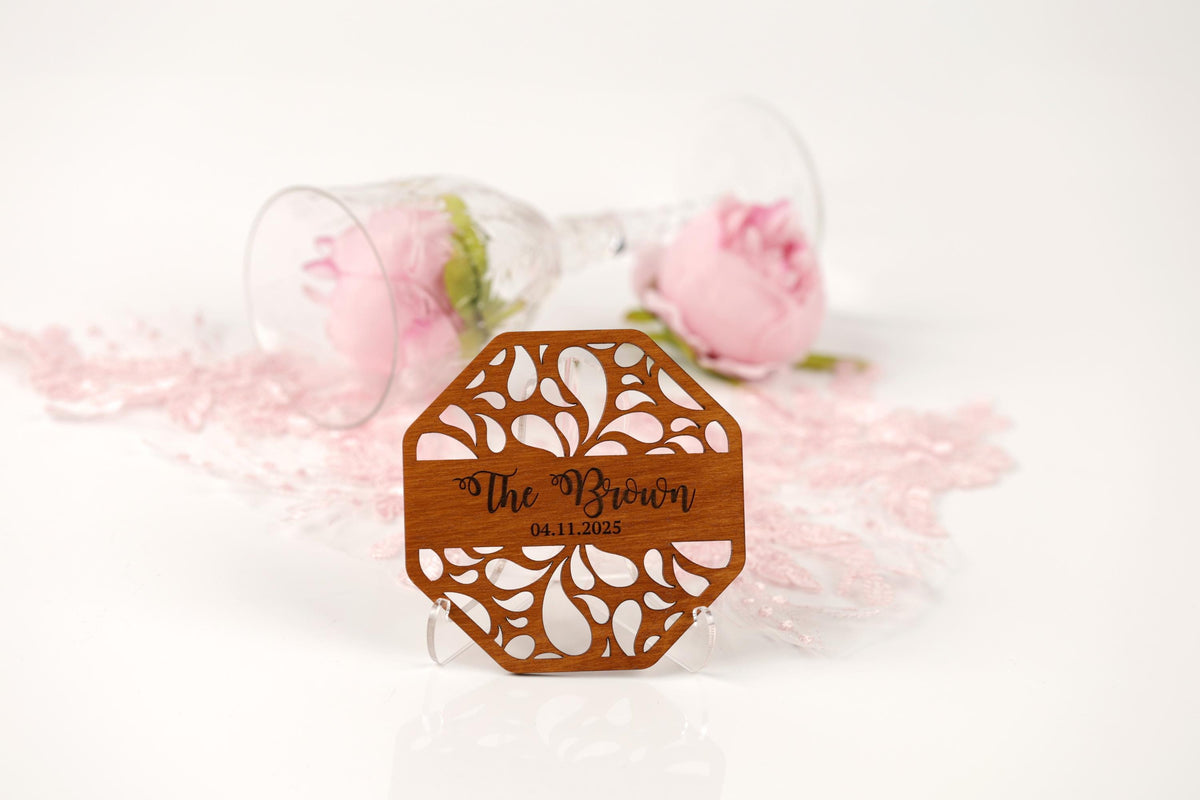 A wooden coaster with a floral design and text, placed on a white surface with a glass of pink roses and pink glitter in the background.