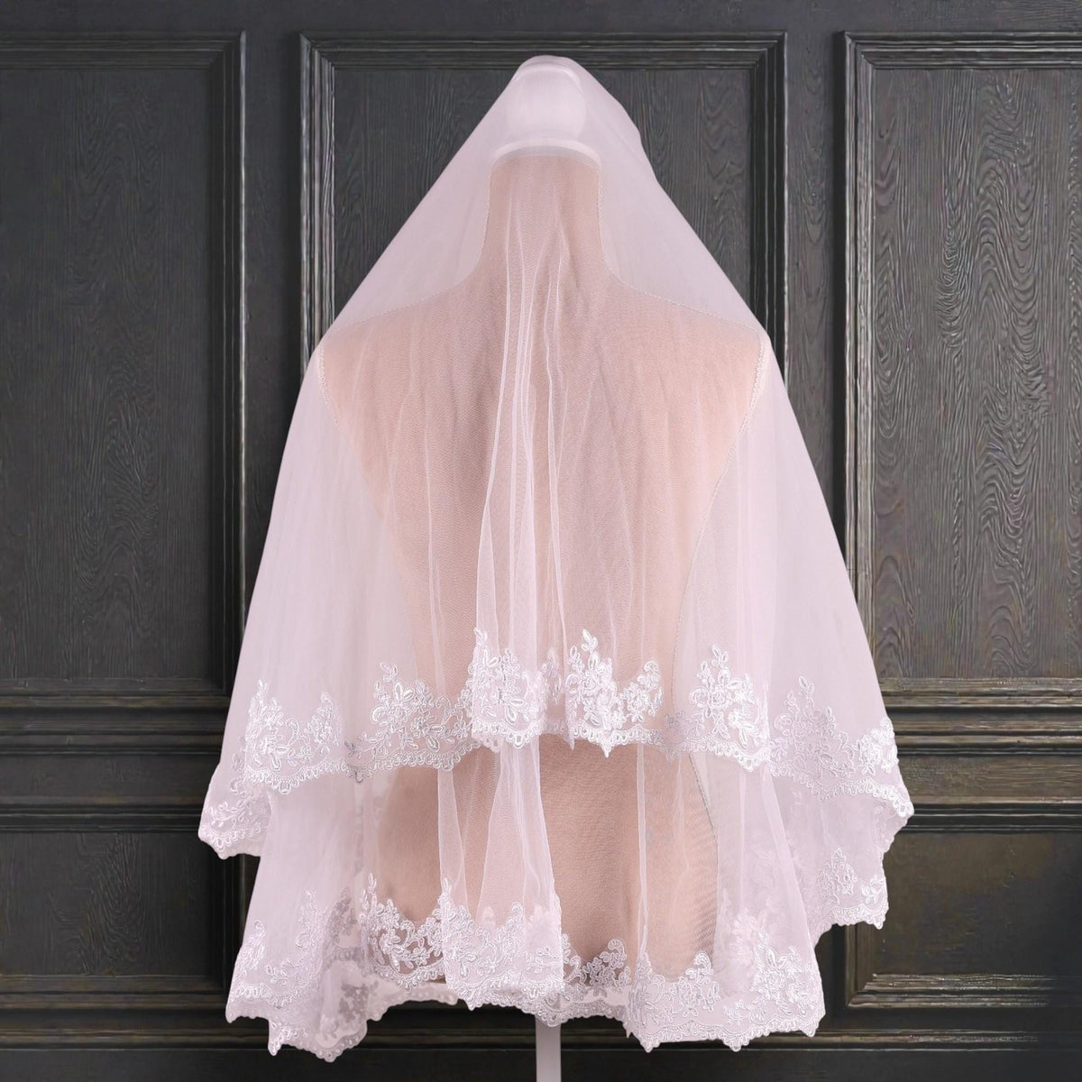 A white wedding veil with lace trim is displayed on a mannequin against a dark background.